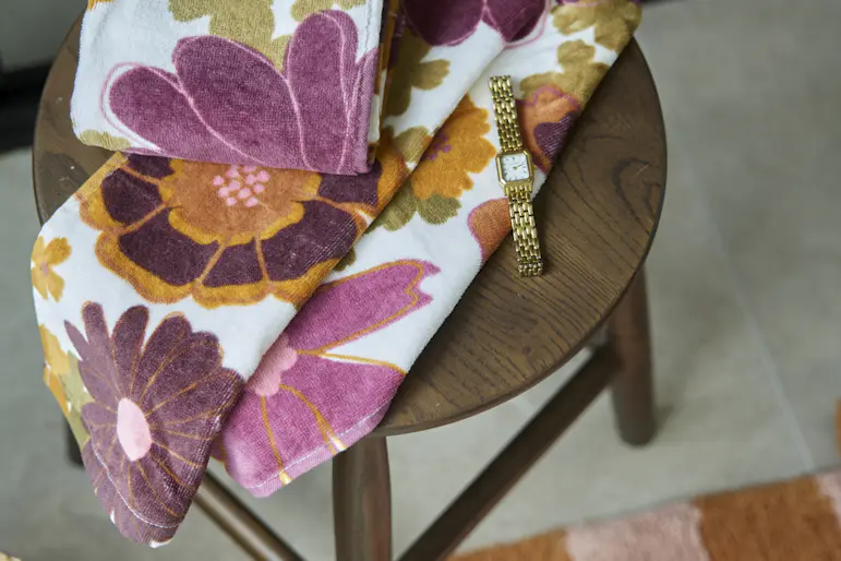 På en brun pall i badrumsmiljö ligger ett stort badlakan och en mindre handduk med blommigt mönser i cerise, organeoch bruna färger.