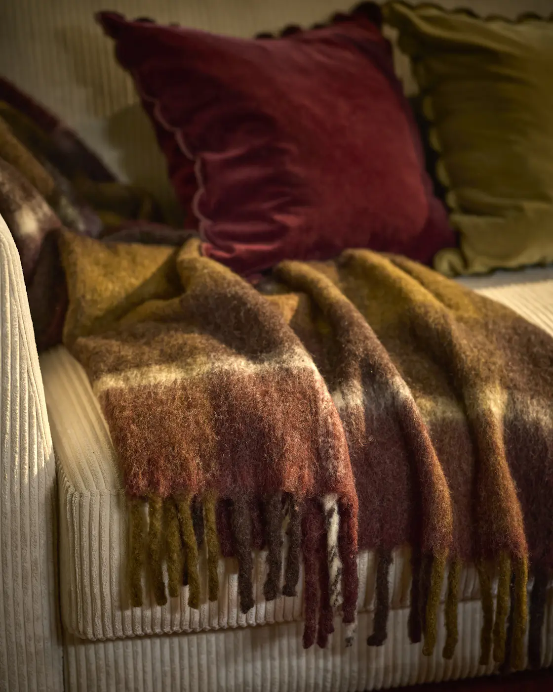 En brun/röd pläd med fransar hänger ner från en beige soffa