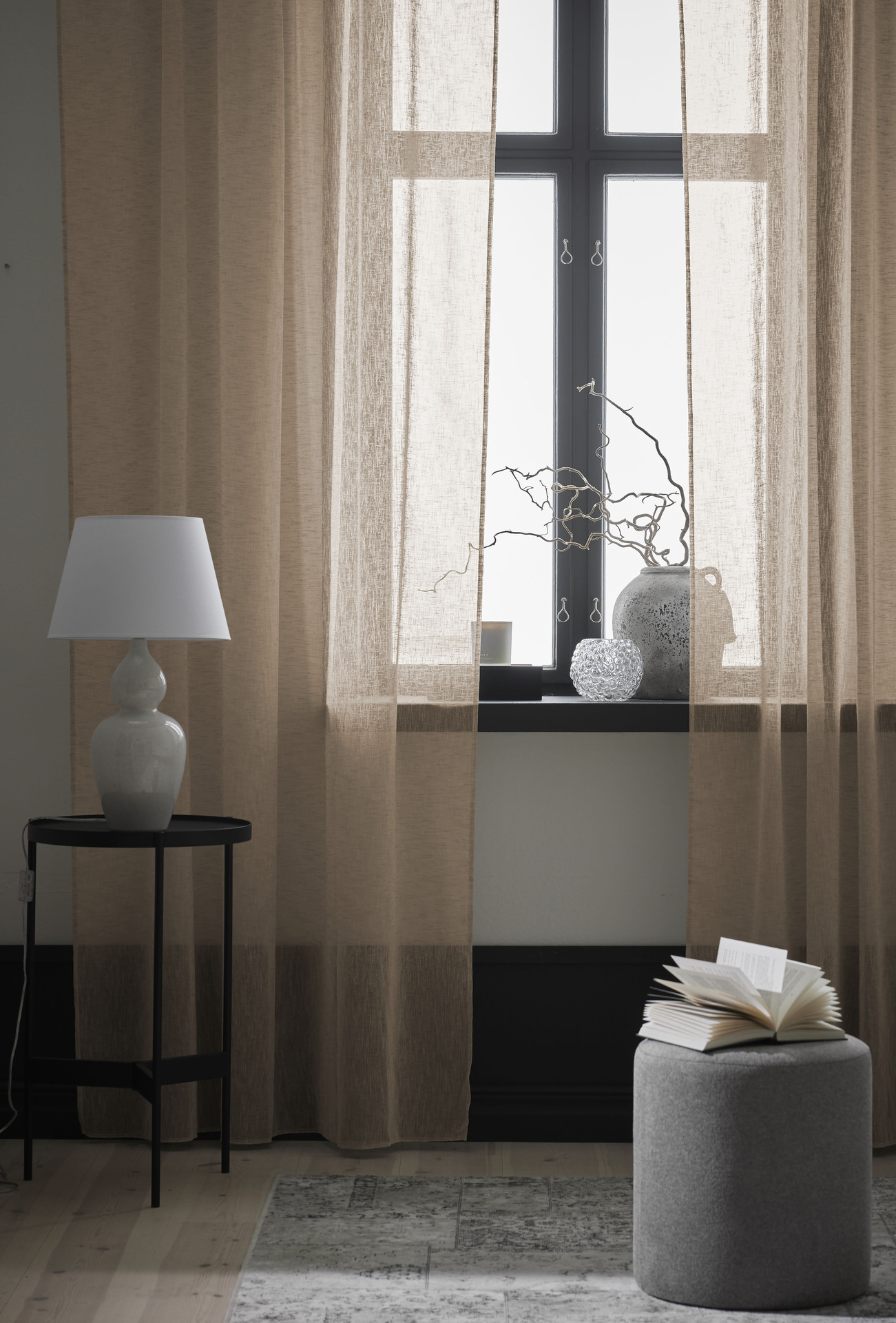 Närbild på en beige gardin med linnestruktur. På golvet står en grå sittpuff och ett svart sidobord med en bordslampa på.