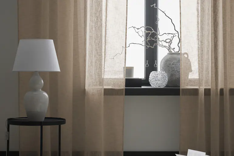 Närbild på en beige gardin med linnestruktur. På golvet står en grå sittpuff och ett svart sidobord med en bordslampa på.