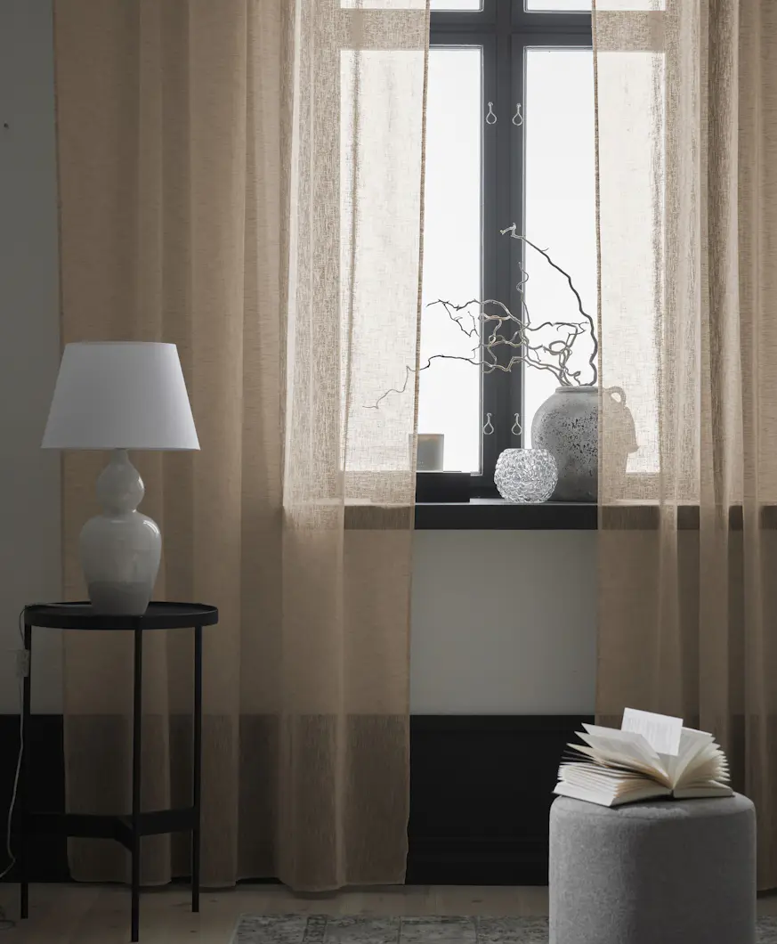 Närbild på en beige gardin med linnestruktur. På golvet står en grå sittpuff och ett svart sidobord med en bordslampa på.