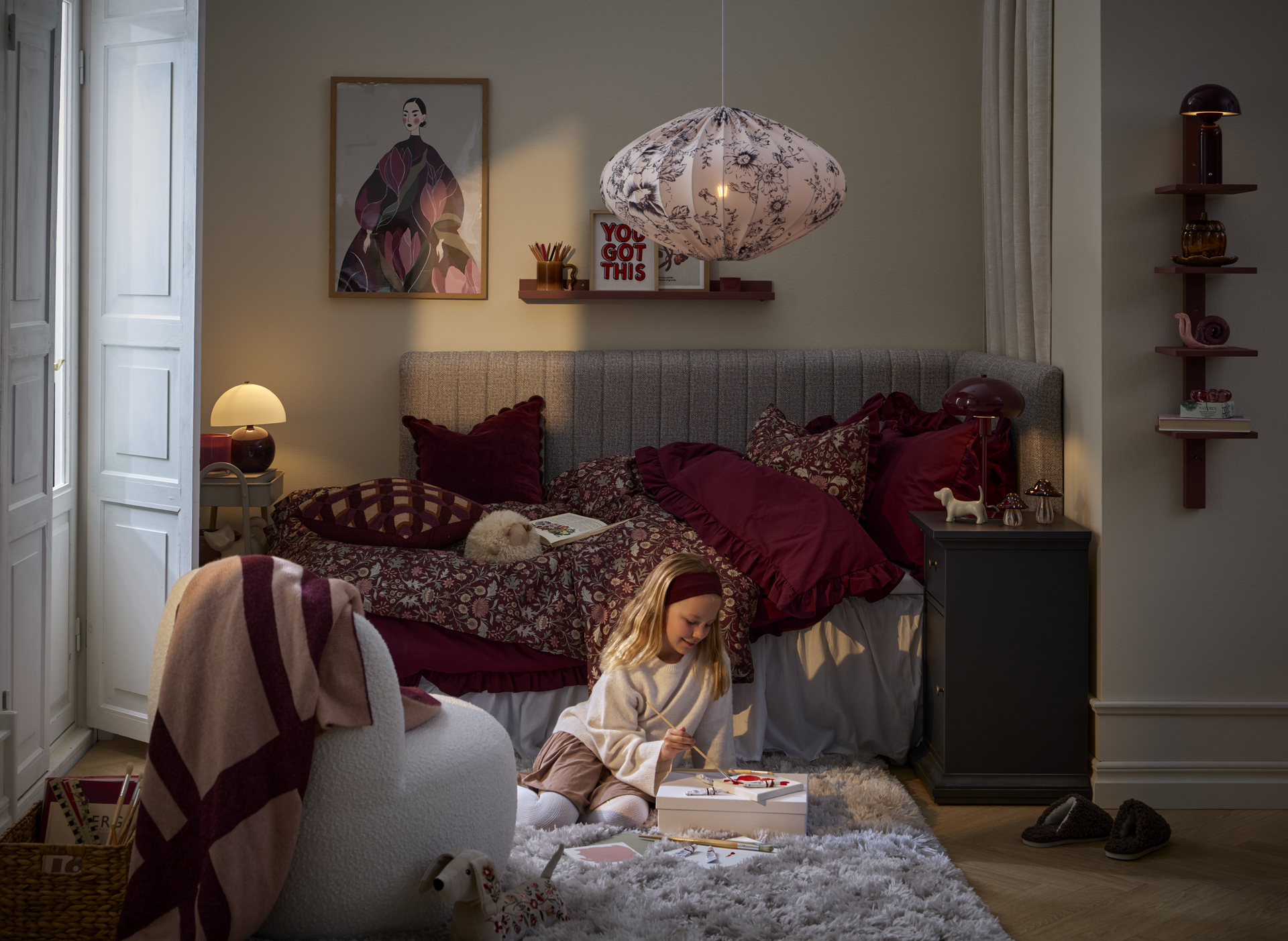 En flicka sitter och målar framför en säng bäddad med vinröda sängkläder.