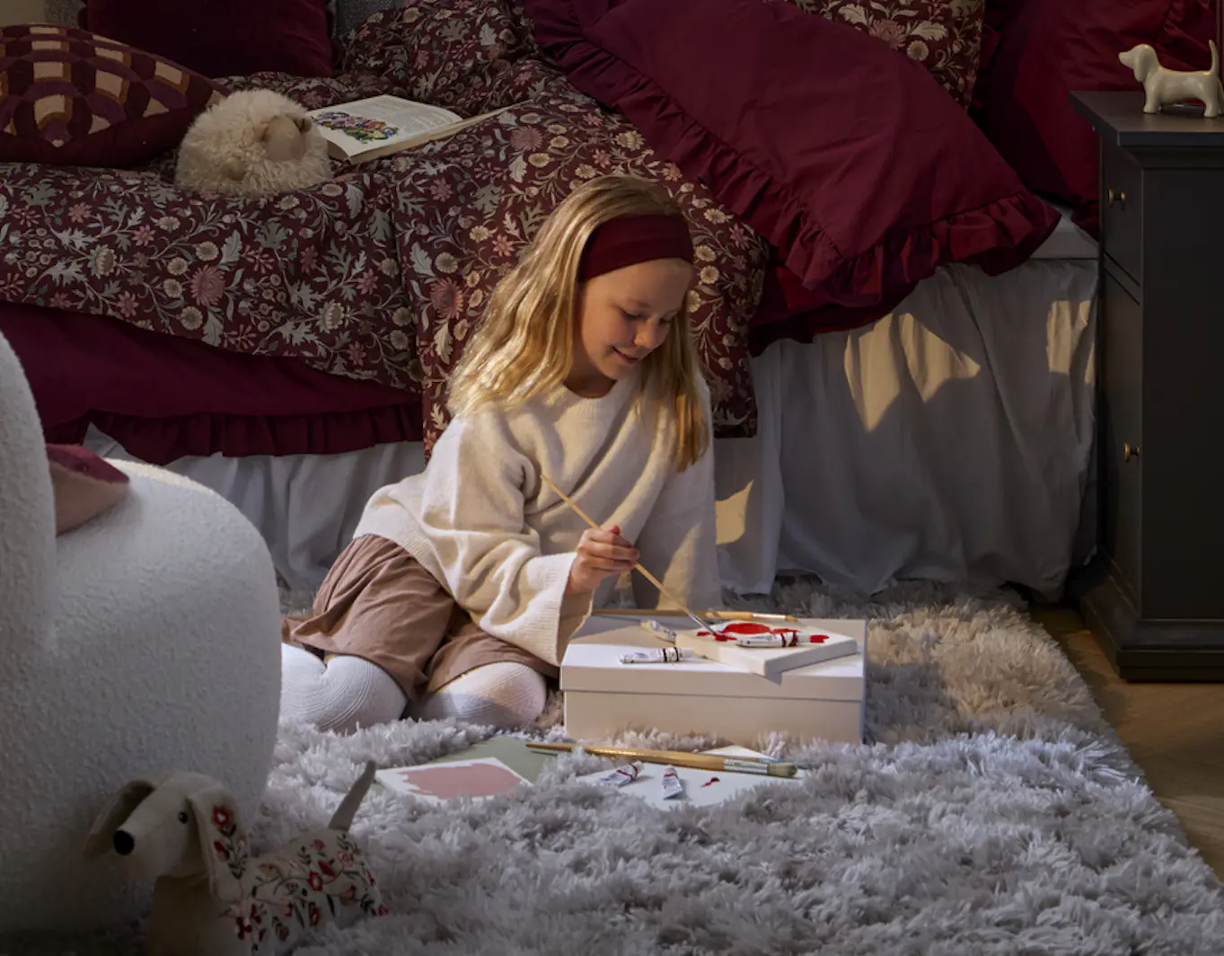 En flicka sitter och målar framför en säng bäddad med vinröda sängkläder.