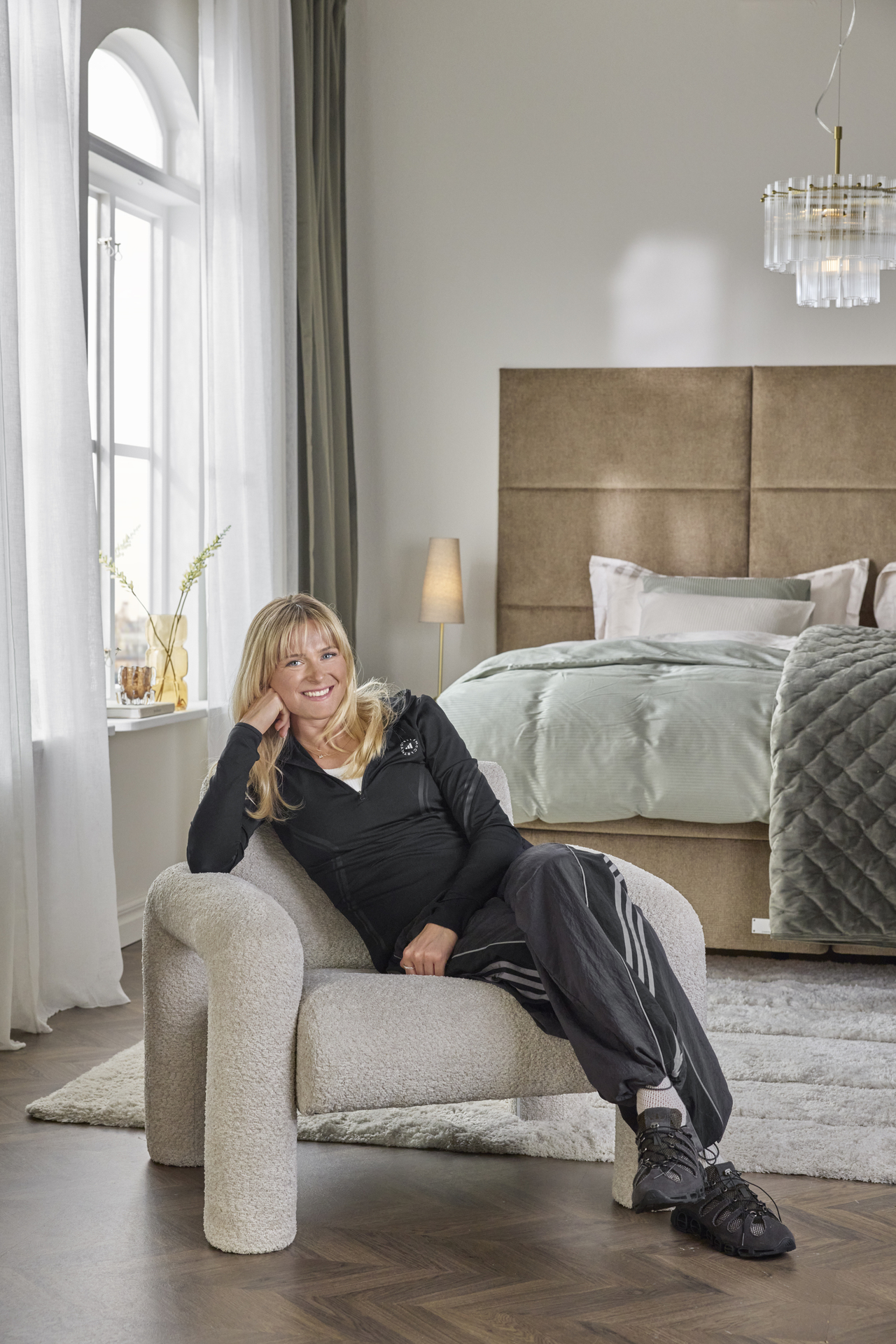Skidåkaren Frida Karlsson iklädd mörka träningskläder sitter i en trendig beige fåtölj i ett ljust sovrum med säng i bakgrunden. 