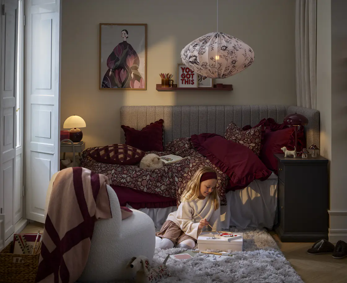 Ett barn sitter på en matta och leker framför en grå säng med röda påslakan