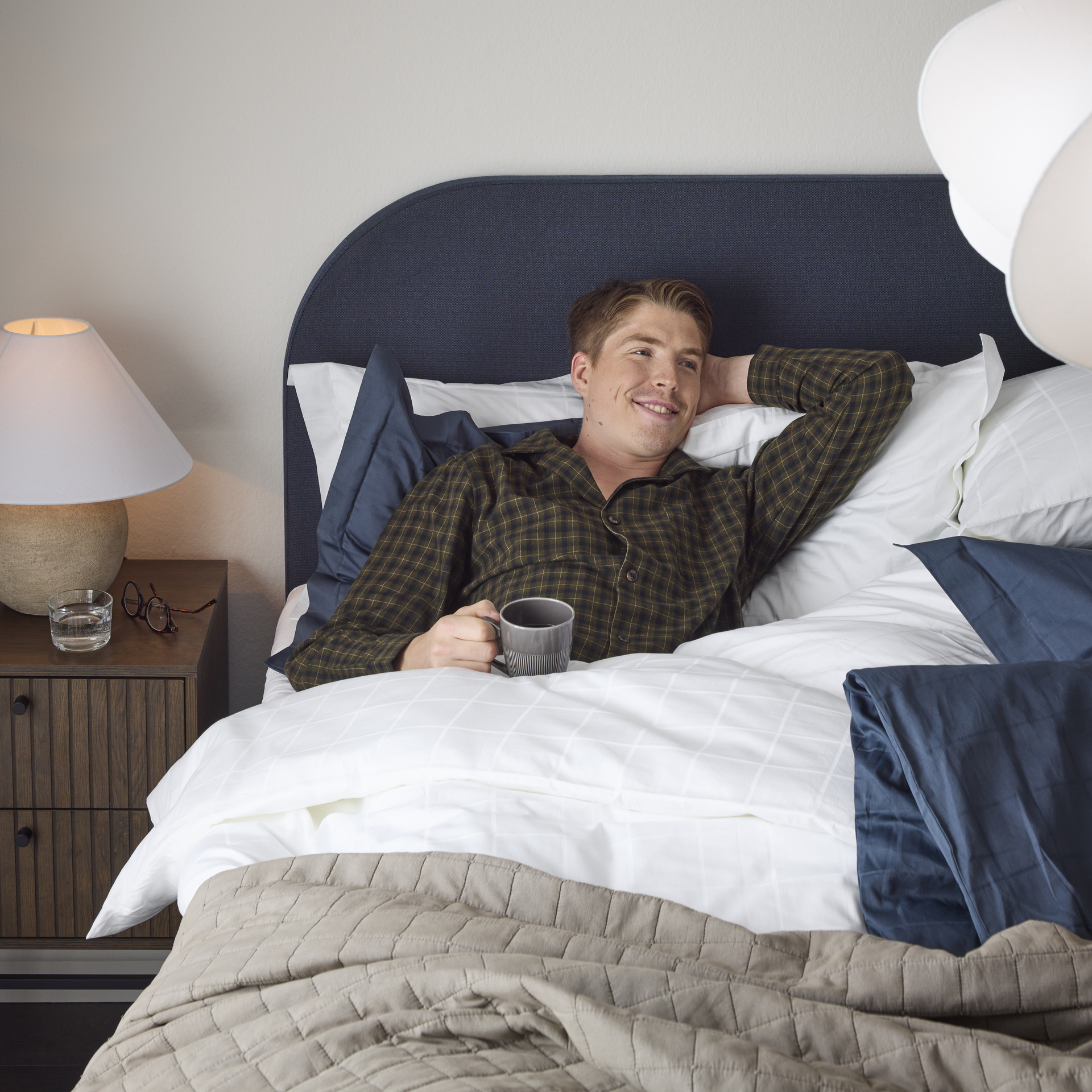 William Poromaa som är längdskidåkare i svenska landslaget ligger i i sängen iklädd mörk grön rutig pyjamas med ena handen bakom nacken vilandes motsänggaveln och i andra handen en kopp kaffe.