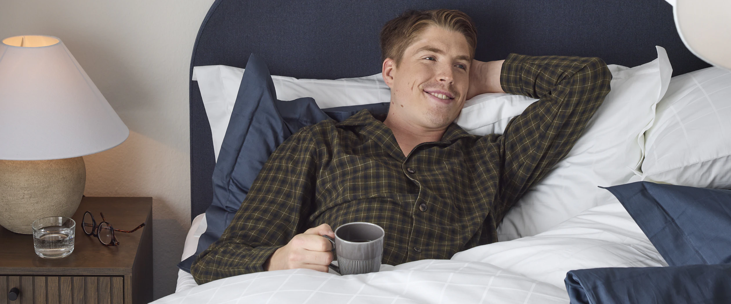 William Poromaa som är längdskidåkare i svenska landslaget ligger i i sängen iklädd mörk grön rutig pyjamas med ena handen bakom nacken vilandes motsänggaveln och i andra handen en kopp kaffe.