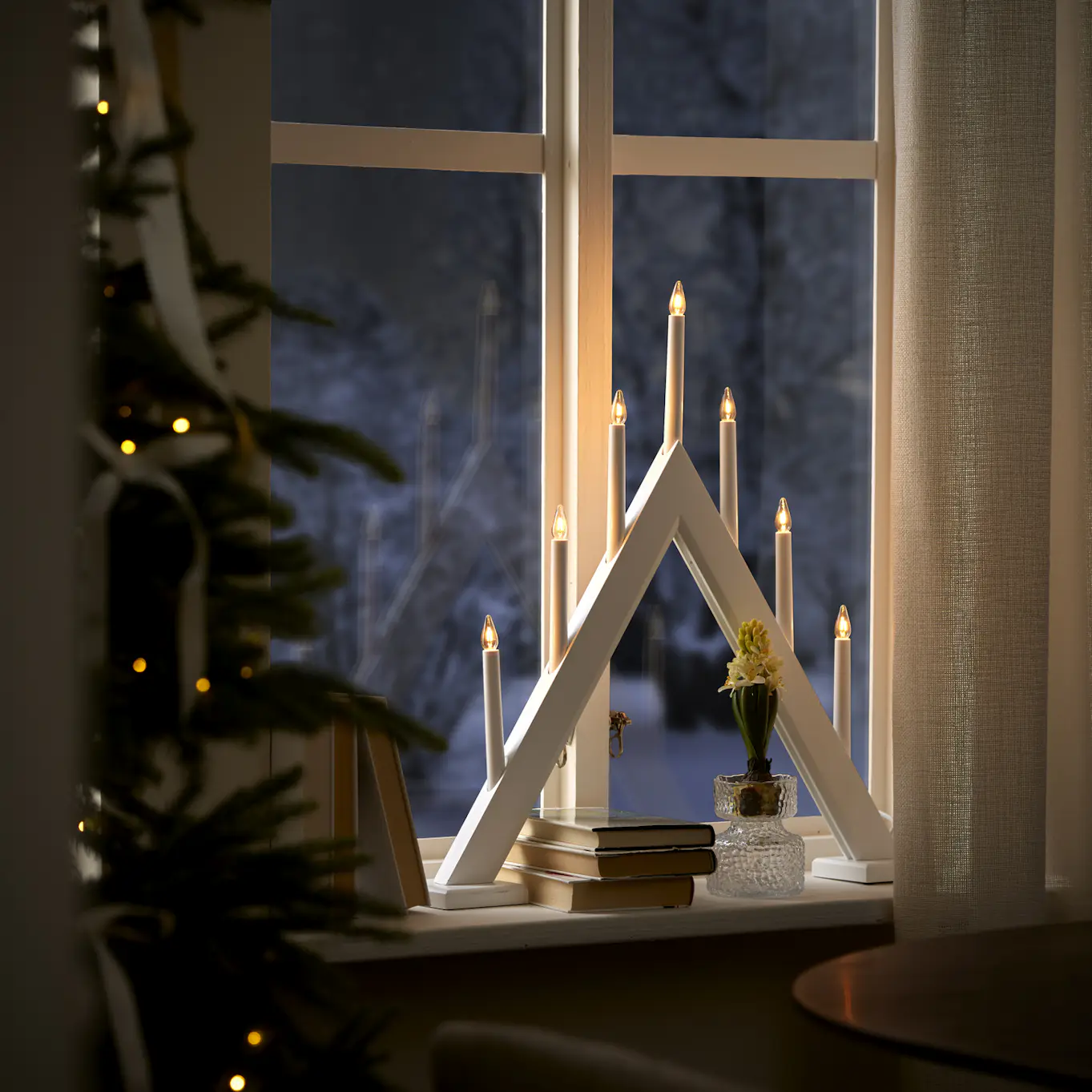I ett fönster står en vit adventsljusstake och en hyacint. Till vänster om fönstret skymtas en julgran.