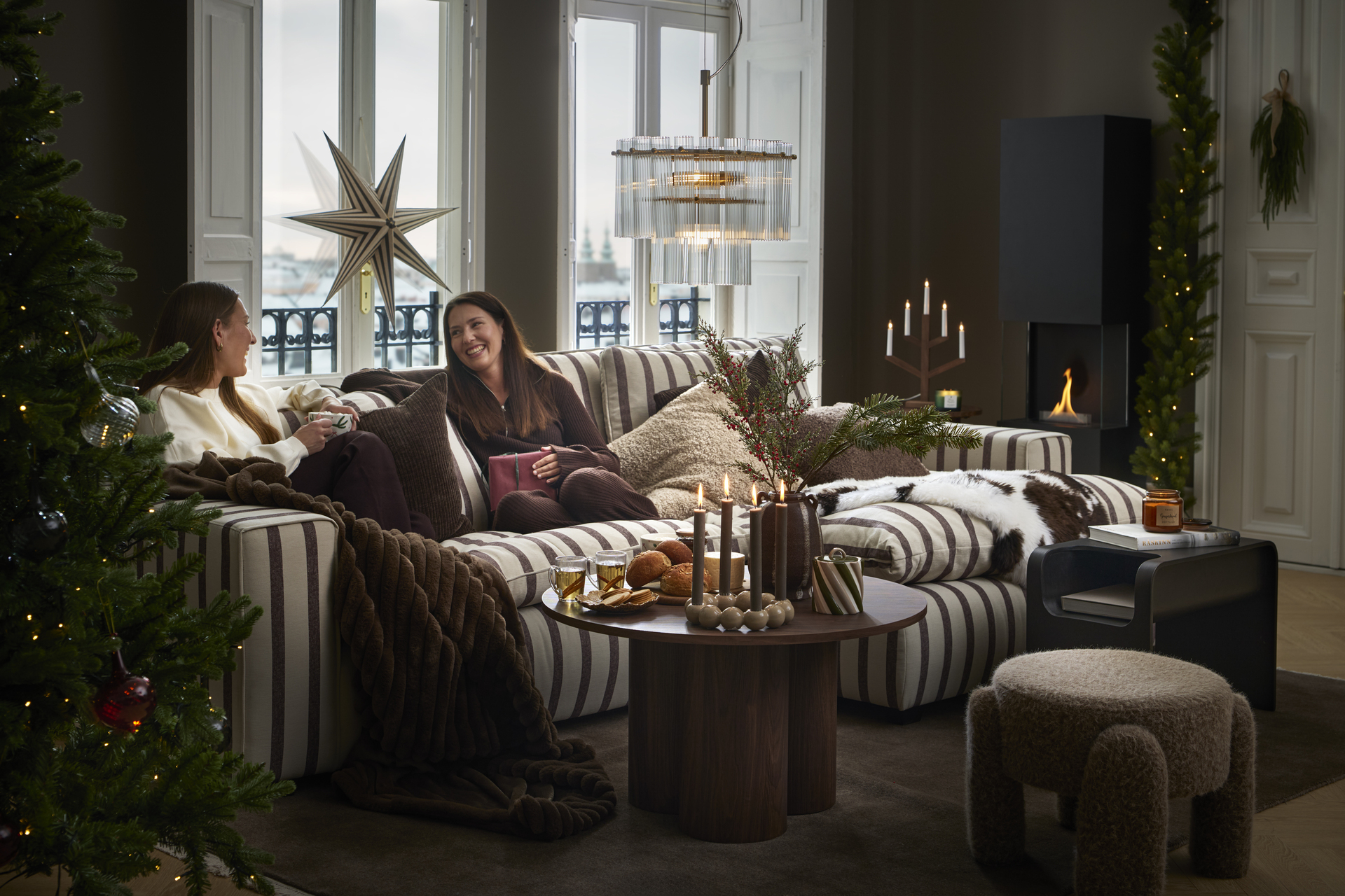 Två kvinnor sitter och samtalar i en  randig soffa som står i ett julpyntat rum intill två fönster