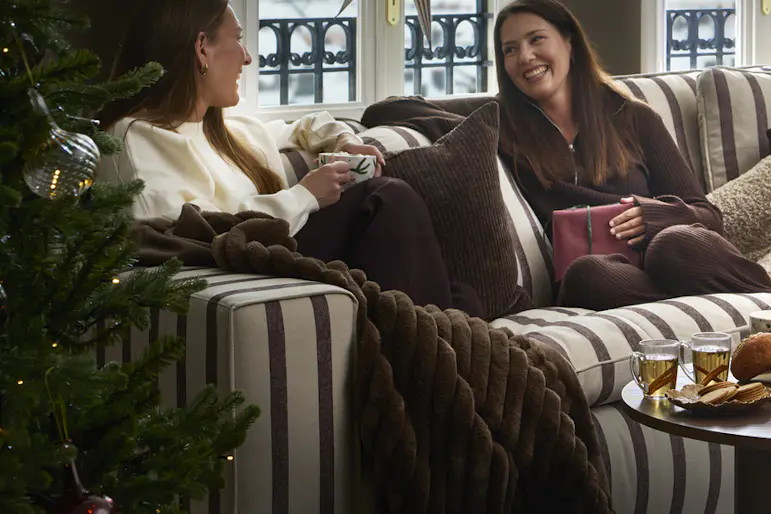 Två kvinnor sitter och samtalar i en randig soffa som står i ett julpyntat rum intill två fönster