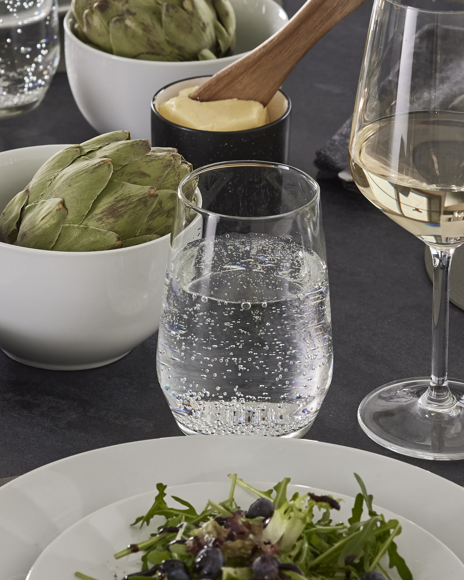 Glas med bubbelvatten står vid ett vinglas, en tallrik med sallad och en skål med kronärtskocka.