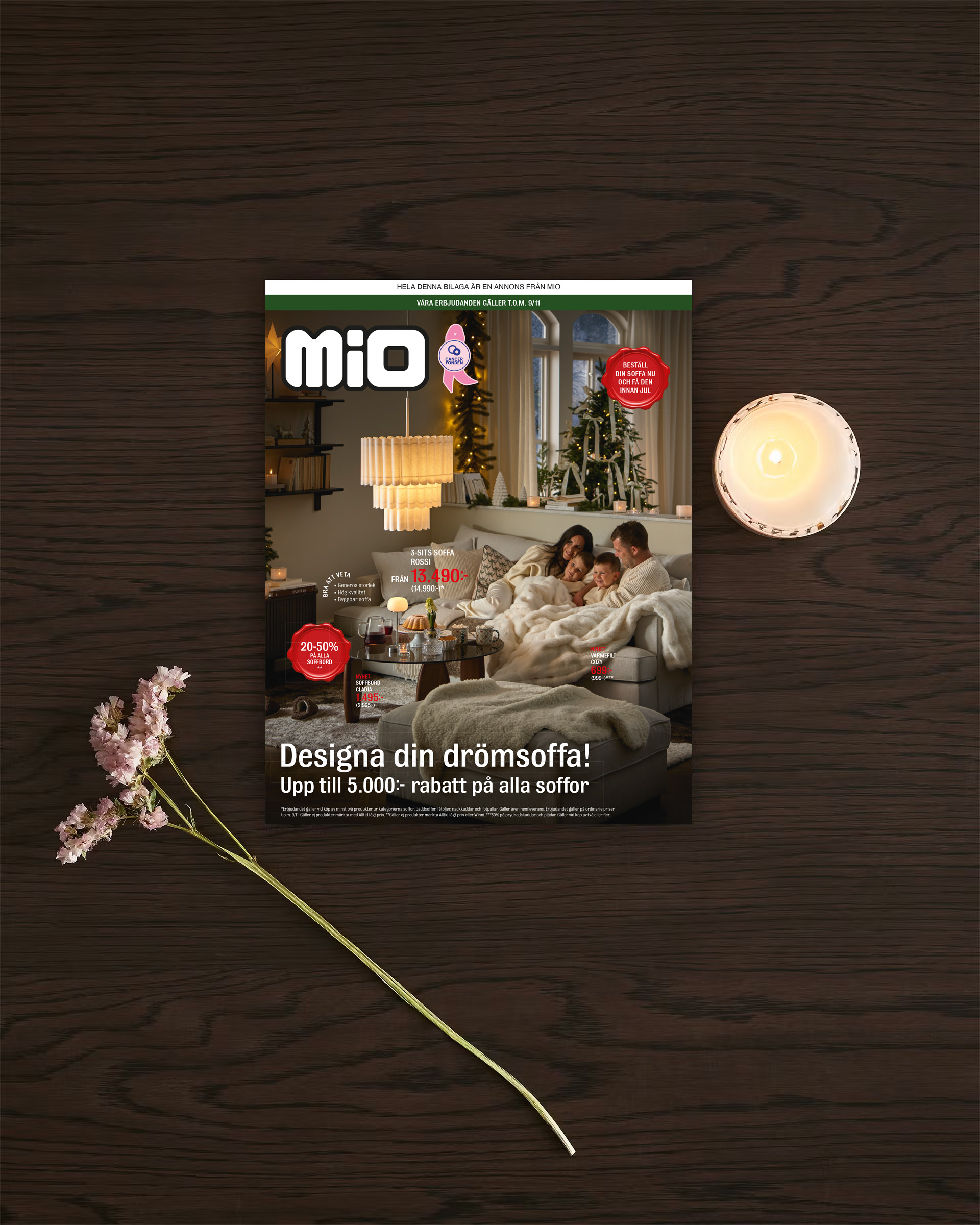 Ett kampanjblad för kampanjen drömsoffa från Mio ligger på ett mörkt träbord med en blomma och ljus bredvid