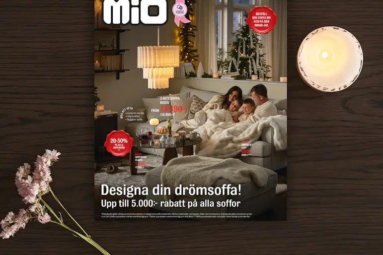 Ett kampanjblad för kampanjen drömsoffa från Mio ligger på ett mörkt träbord med en blomma och ljus bredvid