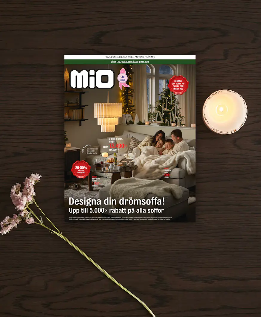 Ett kampanjblad för kampanjen drömsoffa från Mio ligger på ett mörkt träbord med en blomma och ljus bredvid