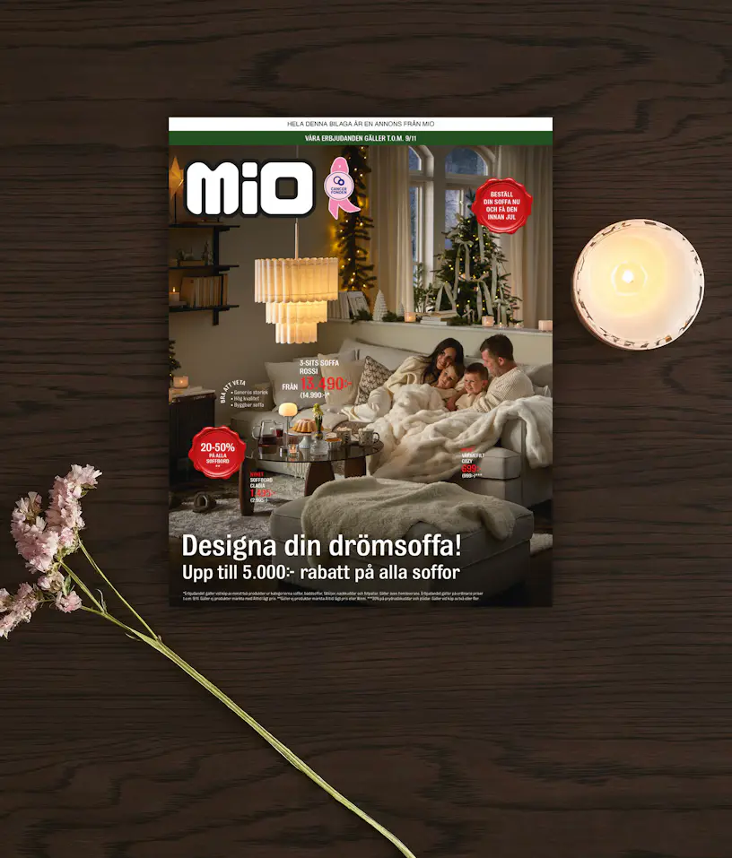 Ett kampanjblad för kampanjen drömsoffa från Mio ligger på ett mörkt träbord med en blomma och ljus bredvid