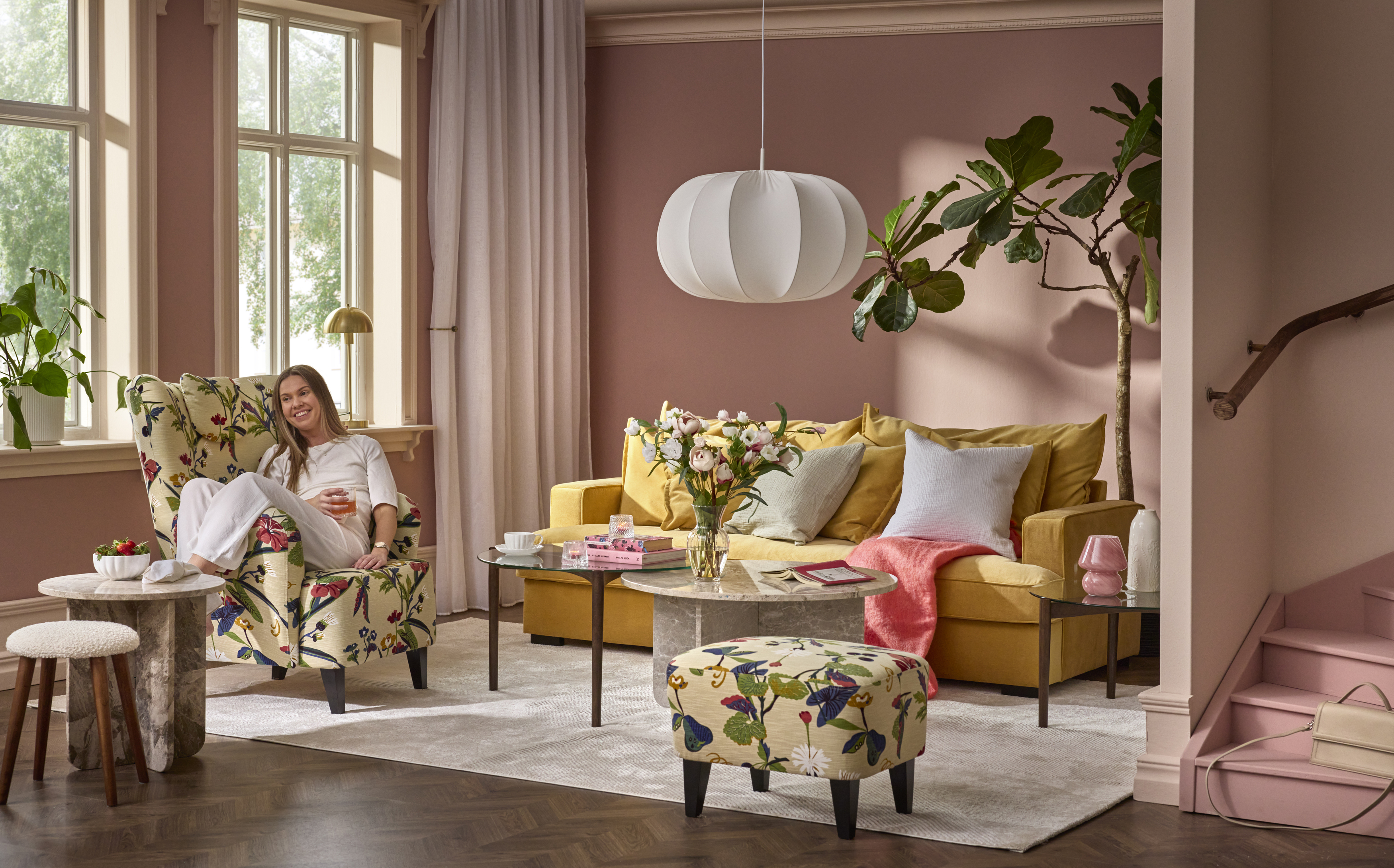 I ett rosa vardagsrum står en gul soffa i sammet, intill soffan sitter en kvinna i en blommig fåtölj