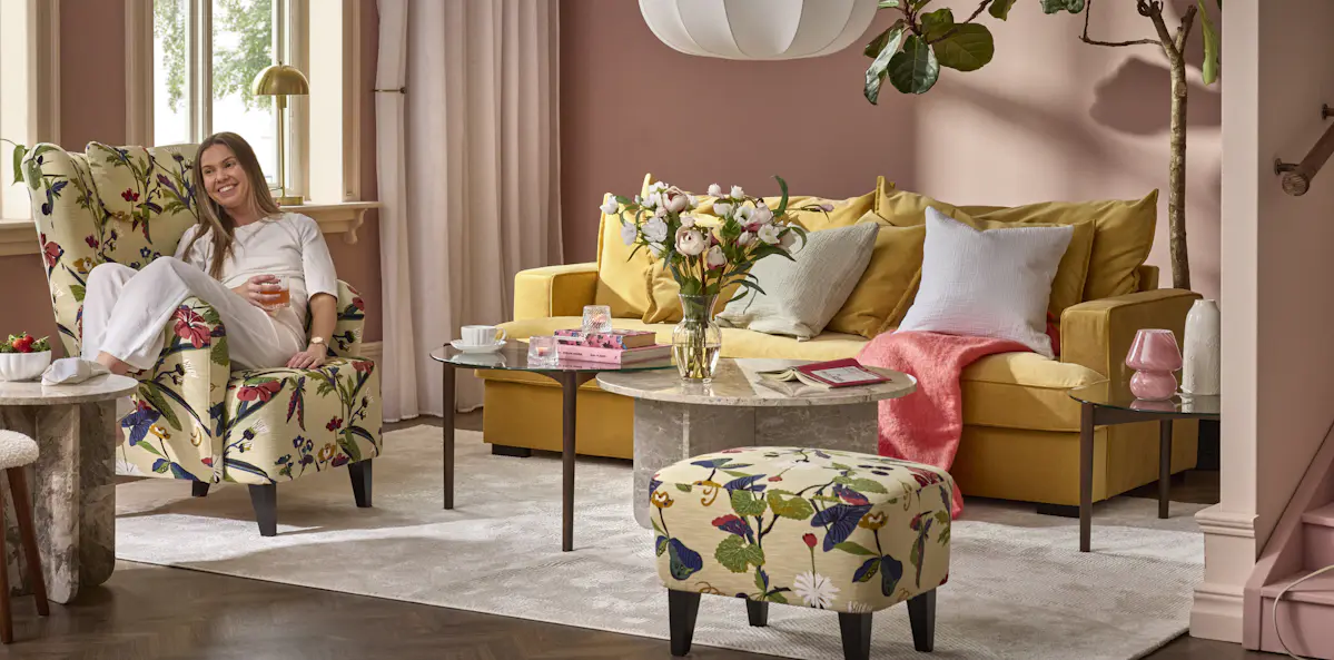 I ett rosa vardagsrum står en gul soffa i sammet, intill soffan sitter en kvinna i en blommig fåtölj