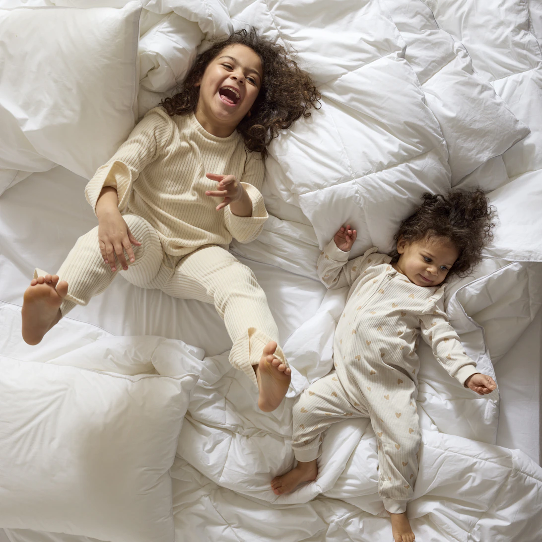 Två små barn busar bland täcken och kuddar i en obäddad säng.