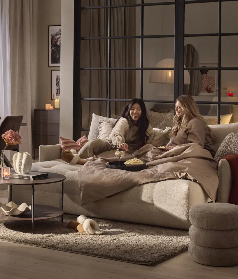 I en vit bäddsoffa sitter två kvinnor under en filt och ler och äter popcorn