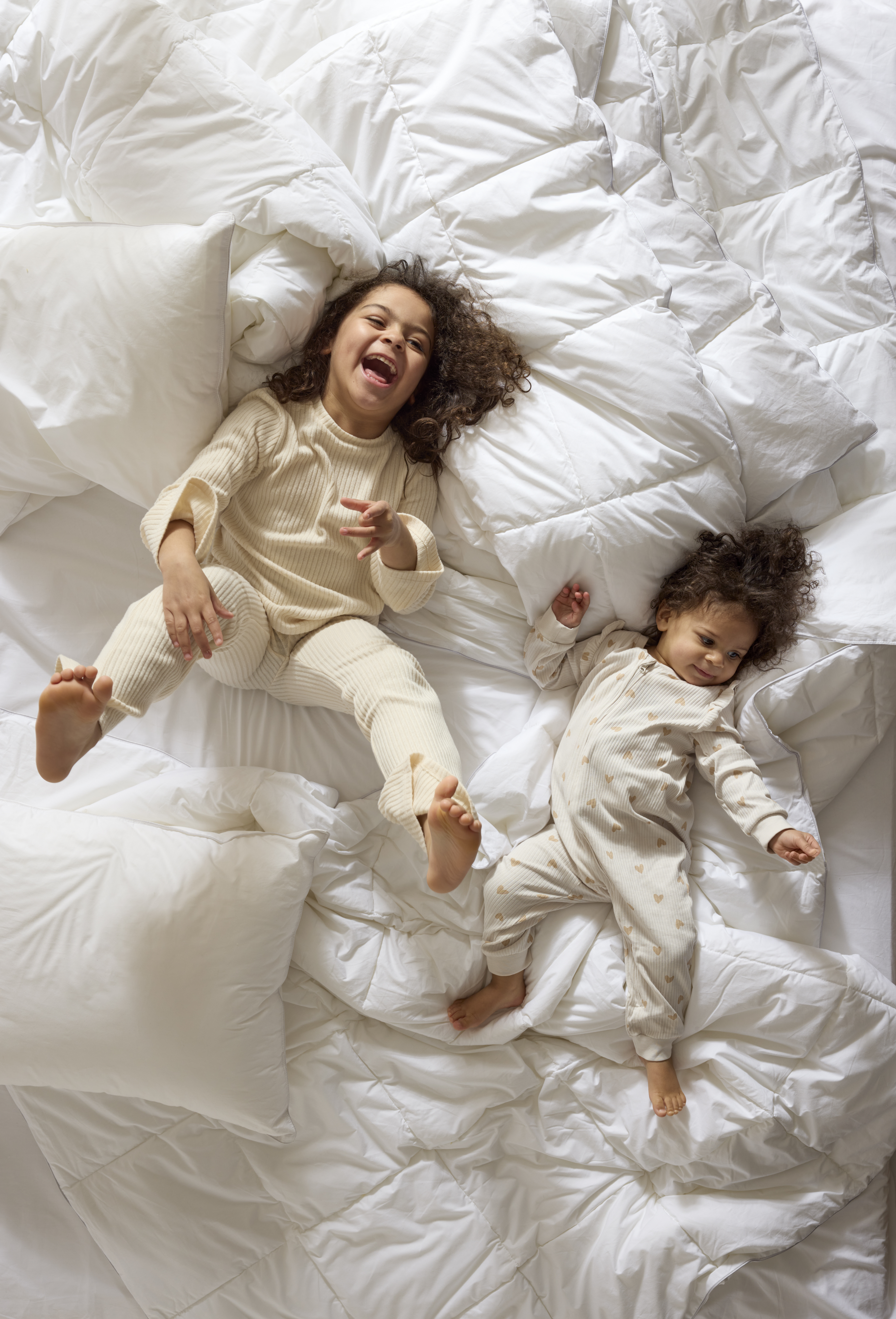 två små barn ligger och skrattar på en säng med täcken och kuddar