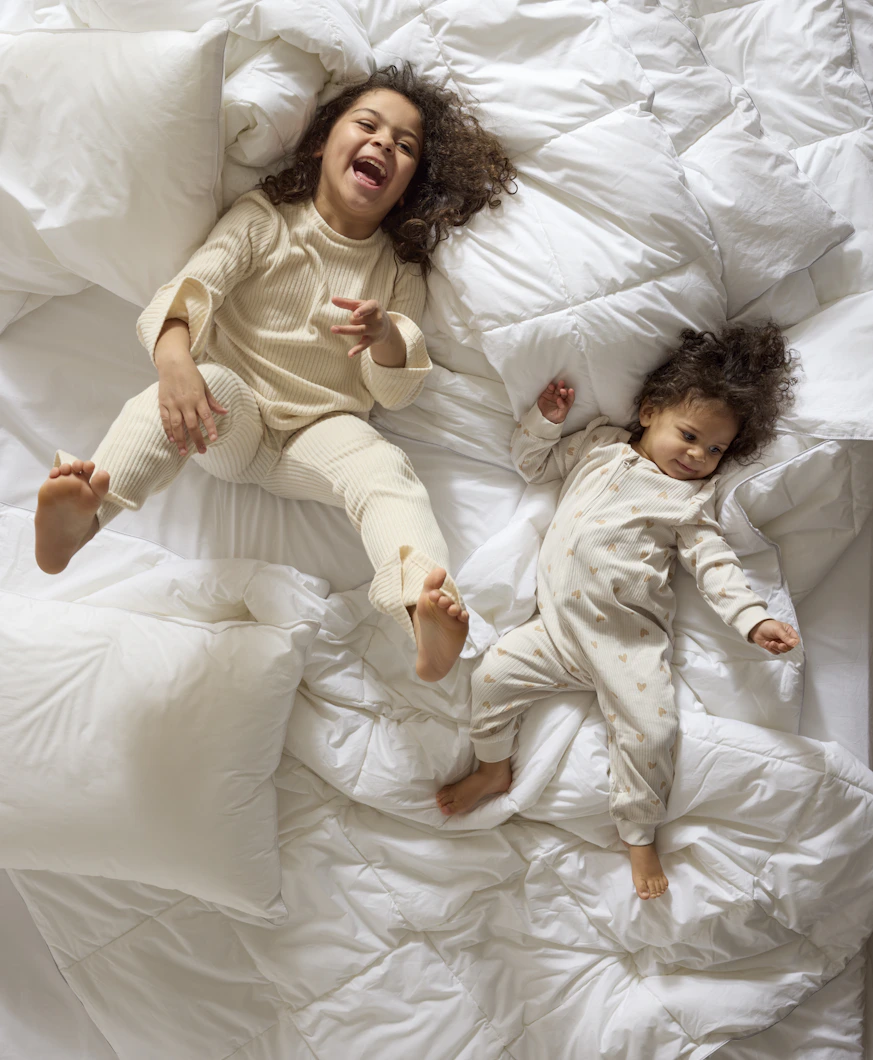 två små barn ligger och skrattar på en säng med täcken och kuddar