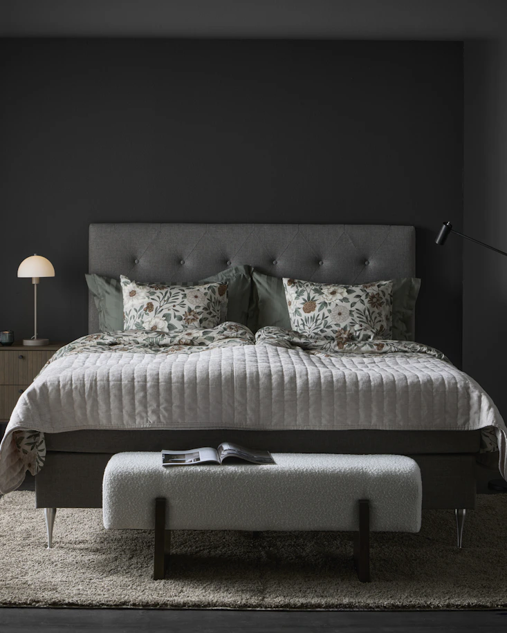 En grå säng med blommigt påslakanset står i ett mörkt rum