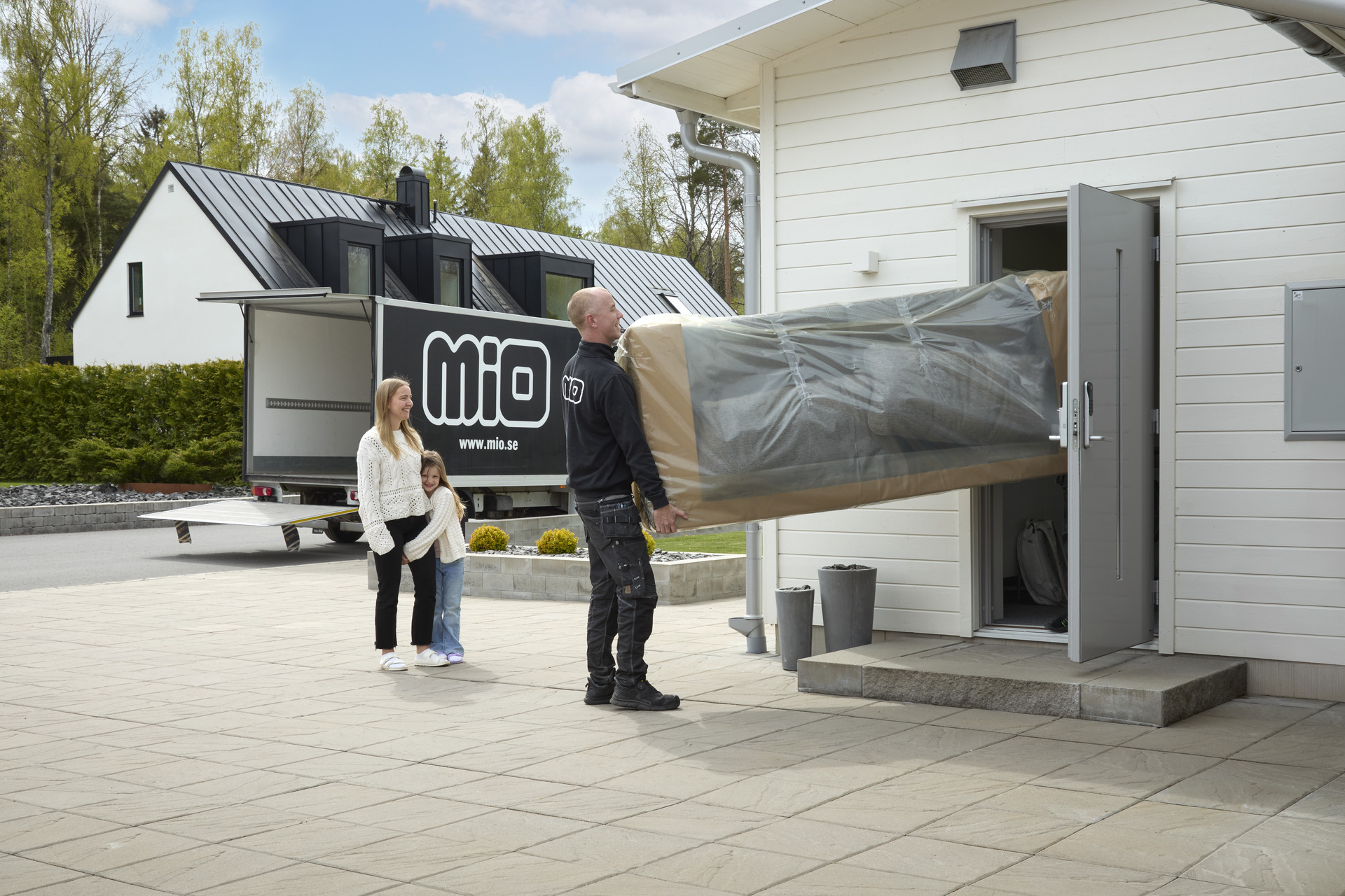 Man med Miologga på ryggen bär in förpackade möbler in i hus, i bakgrunden står en kvinna med litet barn och ser på leendes. 
