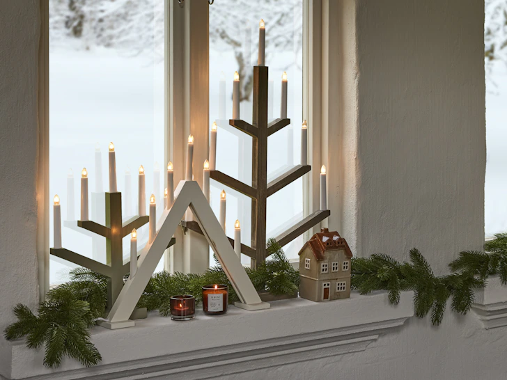 Flera adventsljusstakar i olika färger och höjder ståendes i ett fönster med granris runt