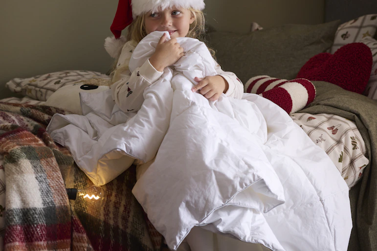 ett barn sittandes på en säng med en tomteluva och kramar ett täcke