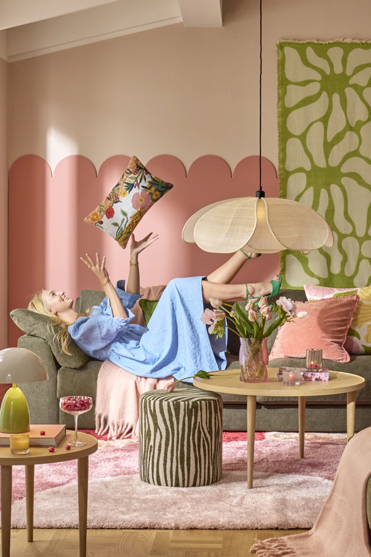 en tjej i blå klänning liggandes på en grön soffa i ett färgglatt rum och kastar upp en kudde i luften