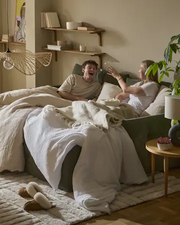 I en grön bäddsoffa sitter en tjej och en kille nedbäddade under ett täcke och kastar popcorn till varandra
