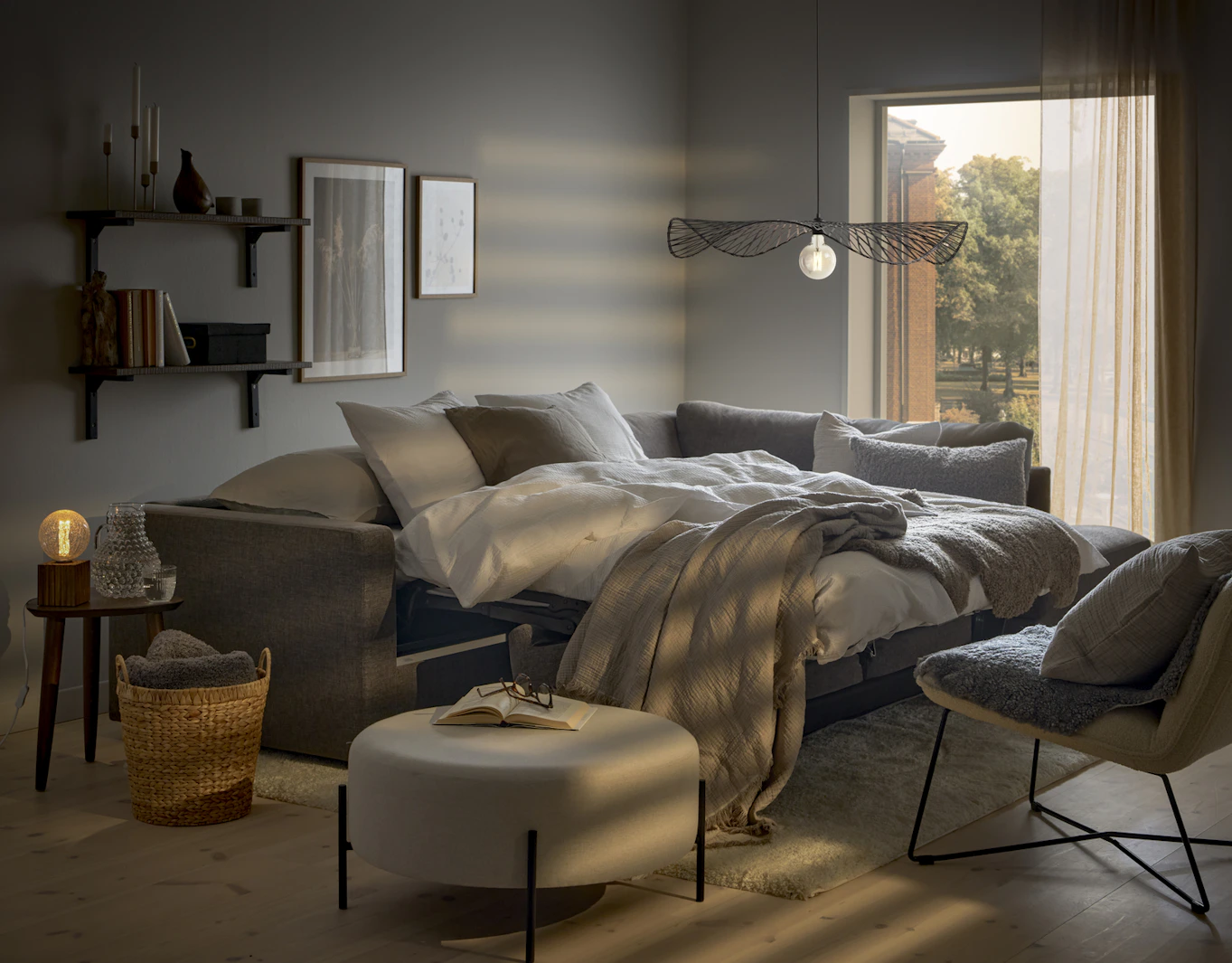 I ett vitt rum står en grå bäddsoffa utbäddad med fluffiga täcken och kuddar