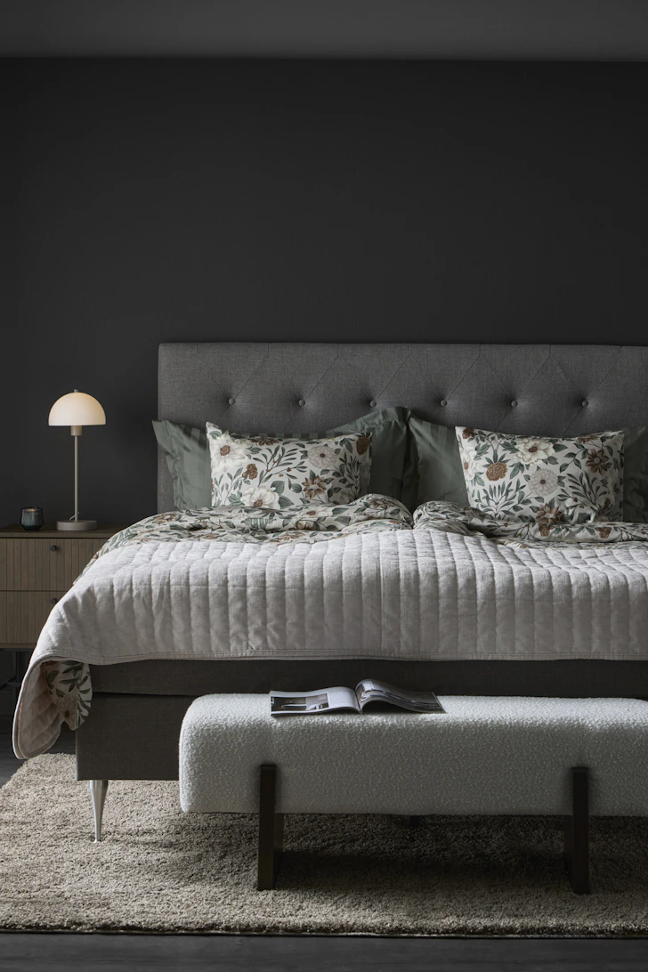 En grå säng med blommigt påslakanset står i ett mörkt rum