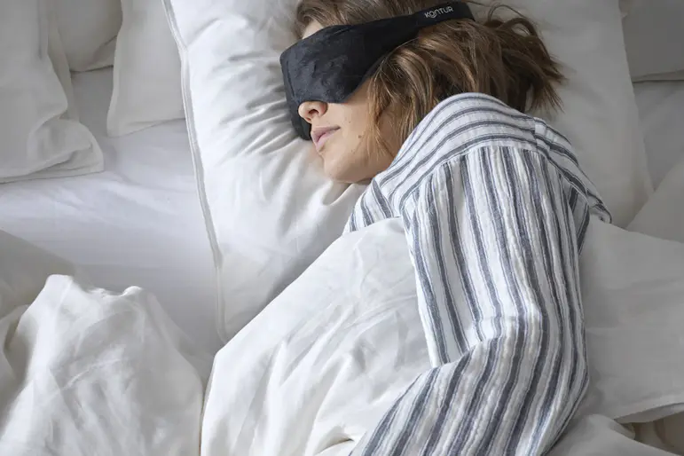 En kvinna i randig pyjamas ligger nedbäddad i en säng med en ögonmask på sig