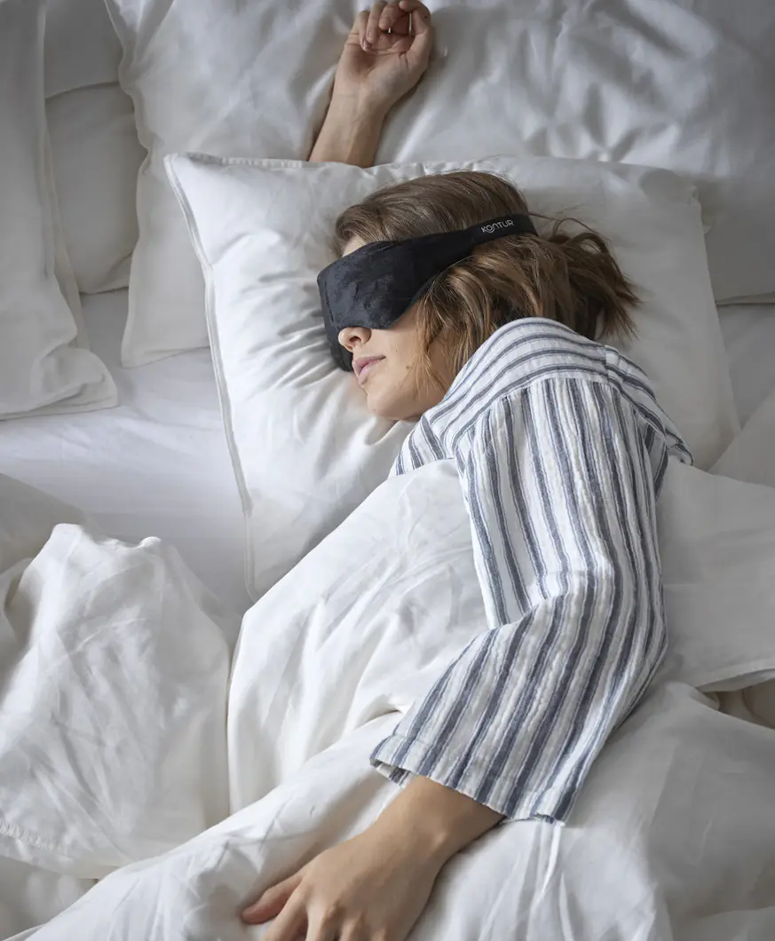 En kvinna i randig pyjamas ligger nedbäddad i en säng med en ögonmask på sig