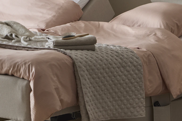 ett grått överkast ligger på en beige säng med rosa sängkläder