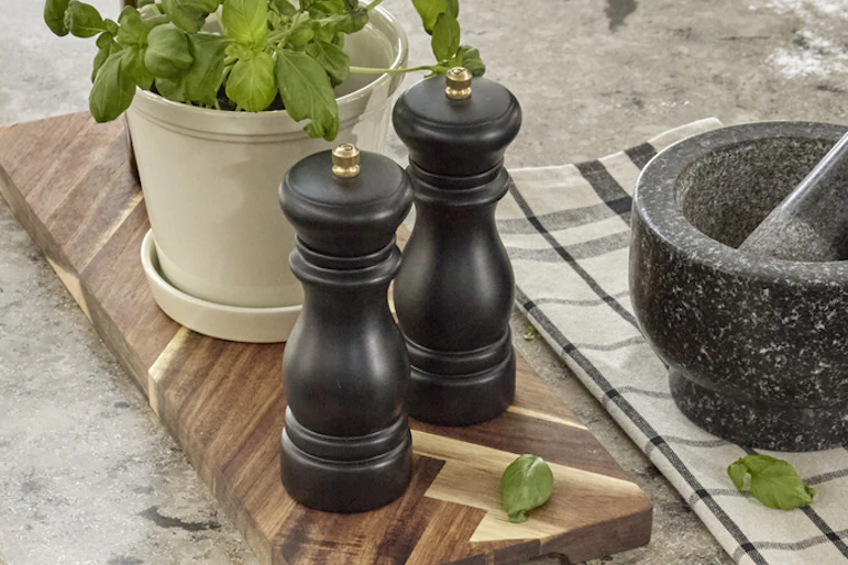 kryddkvarn, olja och basilika i en kruka står på en brun träskärbräda på en köksbänk