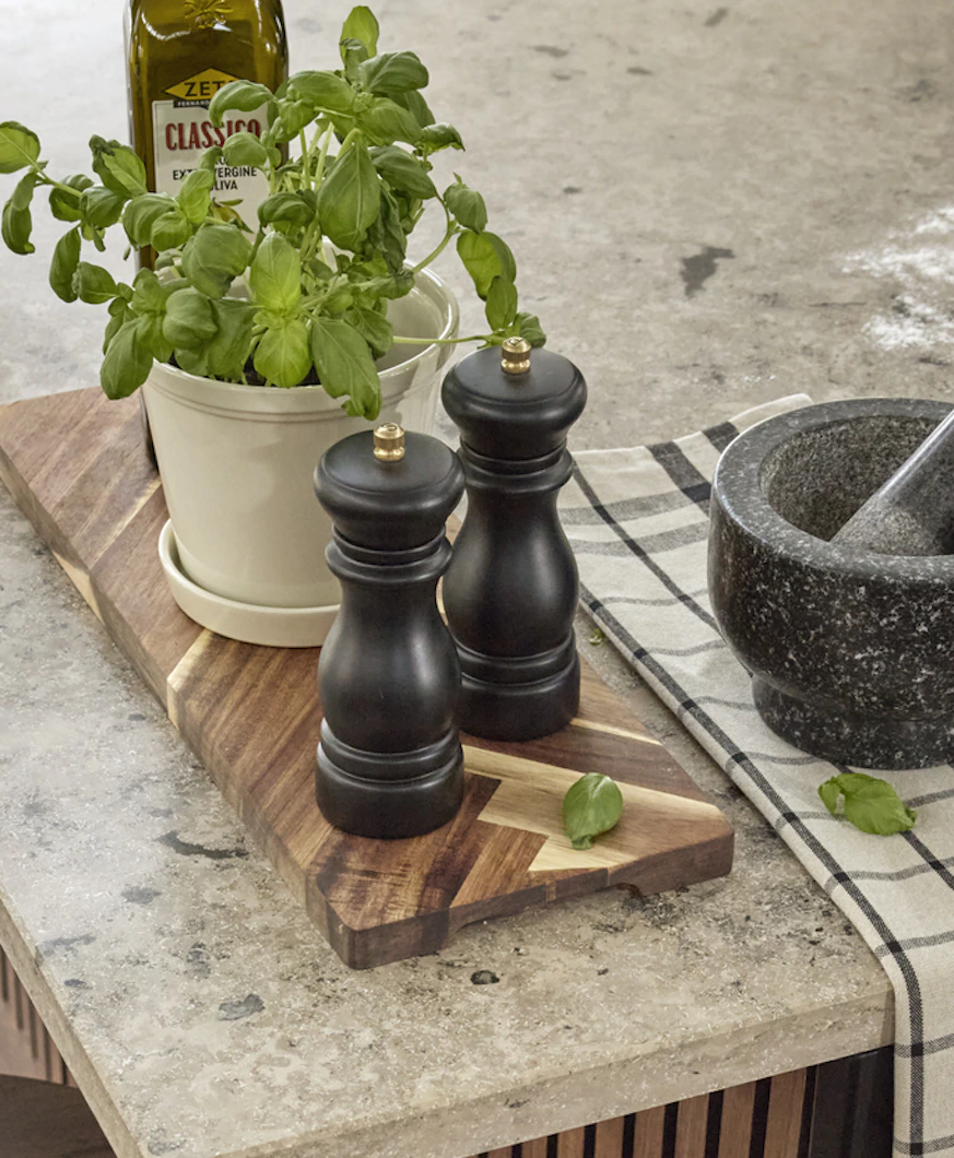 kryddkvarn, olja och basilika i en kruka står på en brun träskärbräda på en köksbänk