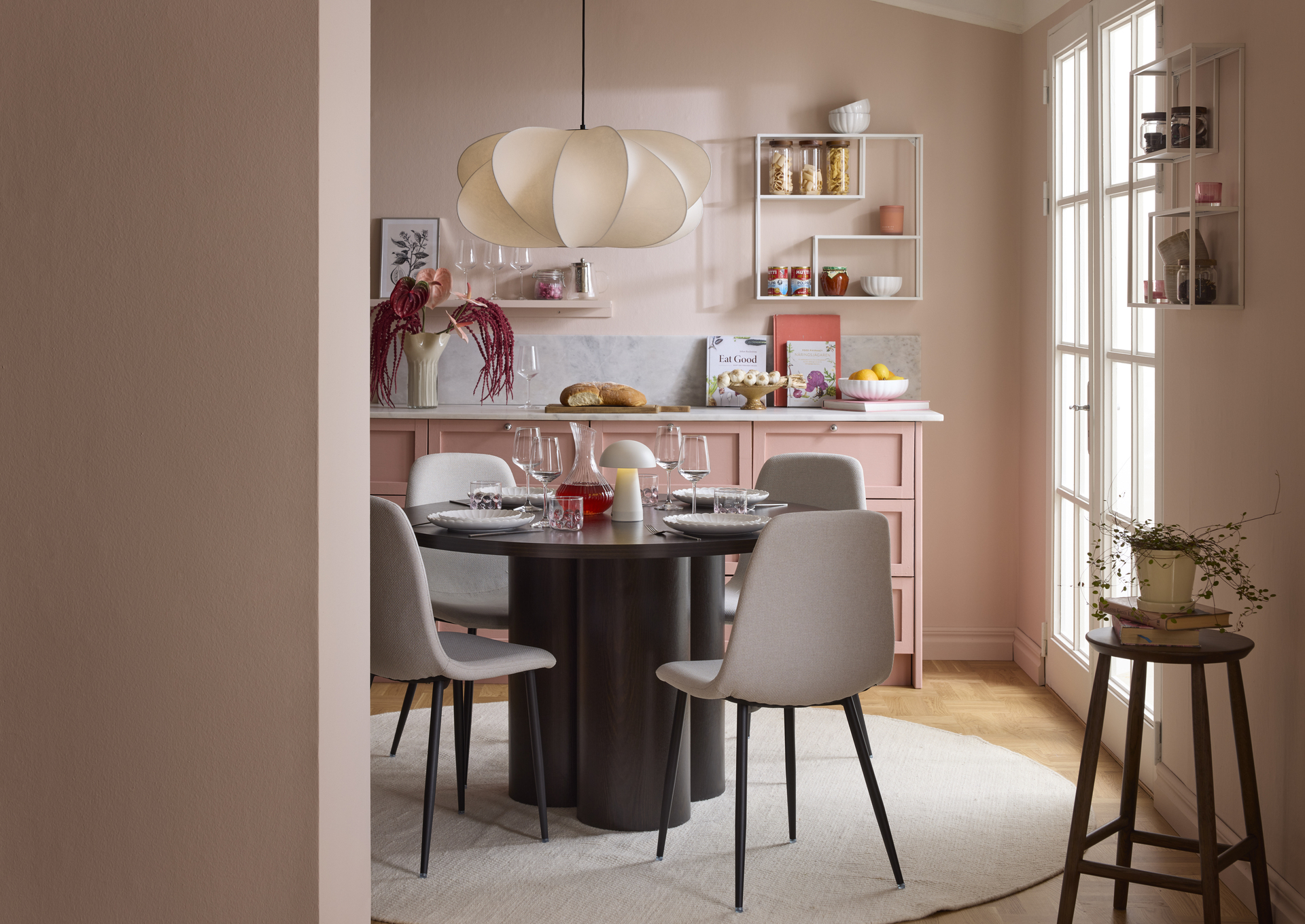 Matbord med kraftig bas av tre cylindrar och rund bordsskiva i mörkt trä samt fyra stolar runtom med ljusbeige klädsel och mörka ben står i ett kök med rosa skåp och väggar.