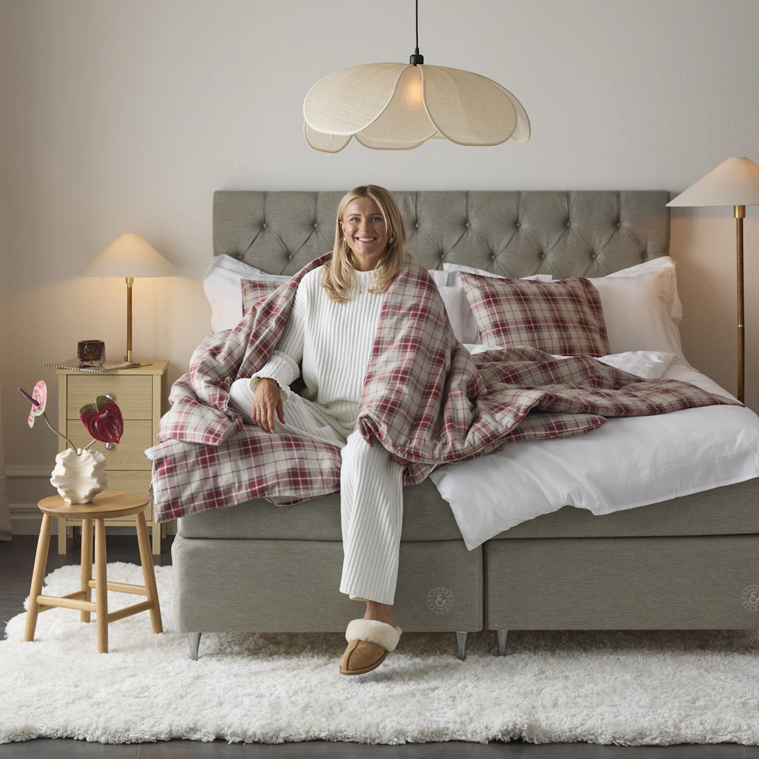 I en grå säng sitter en kvinna inlindad i ett täcke och ler