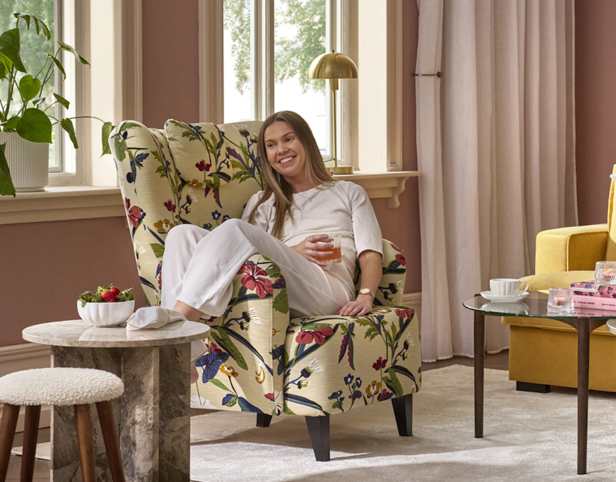 I en mönstrad fåtölj i ett ljusrosa rum sitter en kvinna och ler