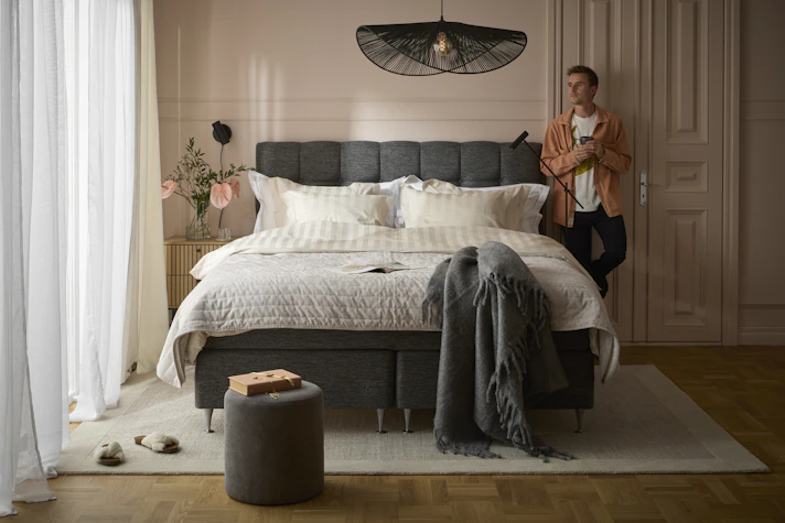 Ung man med skjorta står lutandes med ryggen mot väggen intill sänggavel och blickar ut mot fönster i sovrum.