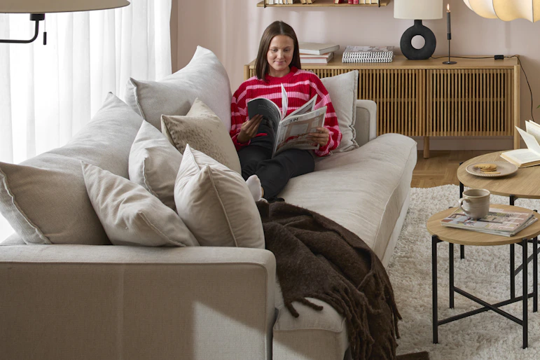 en tjej med rosa tröja sitter i ett vardagsrum där det står en beige soffa, två runda soffbord, en tv-bänk och en vit fåtölj