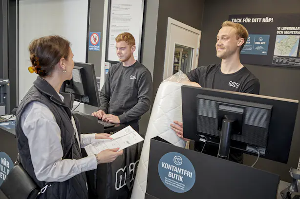 Manlig butiksmedarbetare pratar leende med kund vid kassadisken.