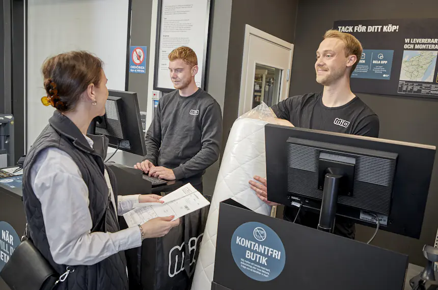 Manlig butiksmedarbetare pratar leende med kund vid kassadisken.