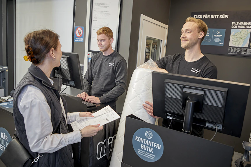 Manlig butiksmedarbetare pratar leende med kund vid kassadisken.