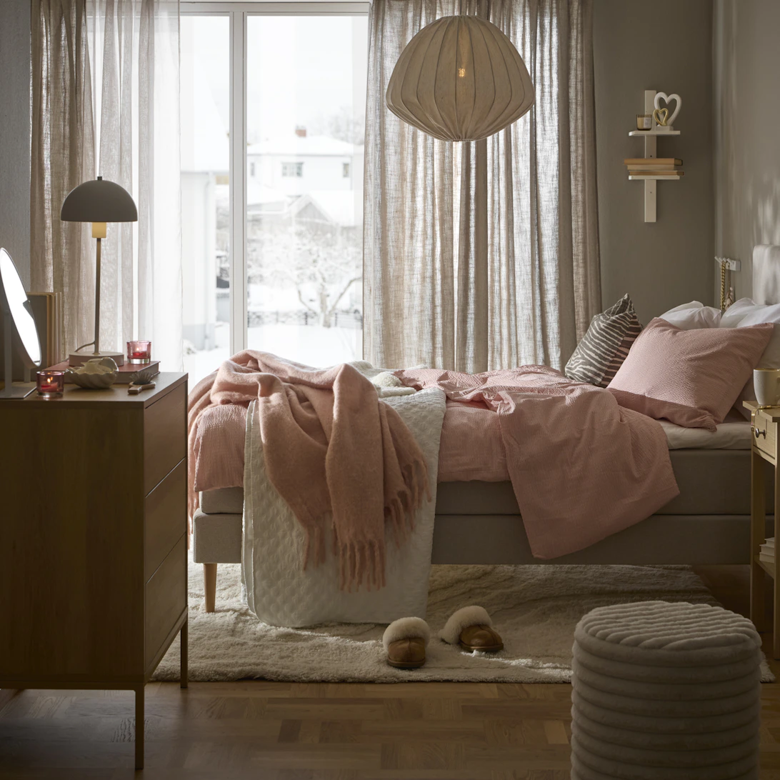 ljusgrå mindre säng står i ett skandinaviskt sovrum med ljust rosa påslakanset liggandes över sängen.