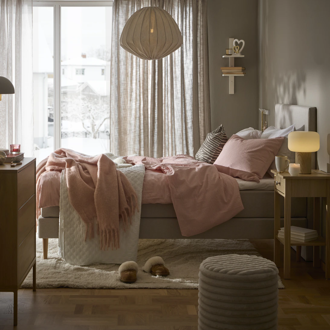 ljusgrå mindre säng står i ett skandinaviskt sovrum med ljust rosa påslakanset liggandes över sängen.
