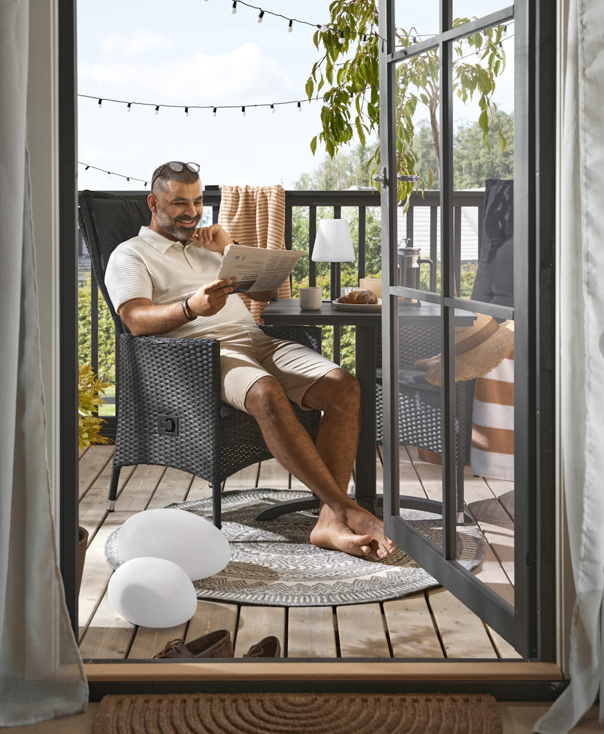 PÅ en balkong sitter en man i en stol och läser tidningen