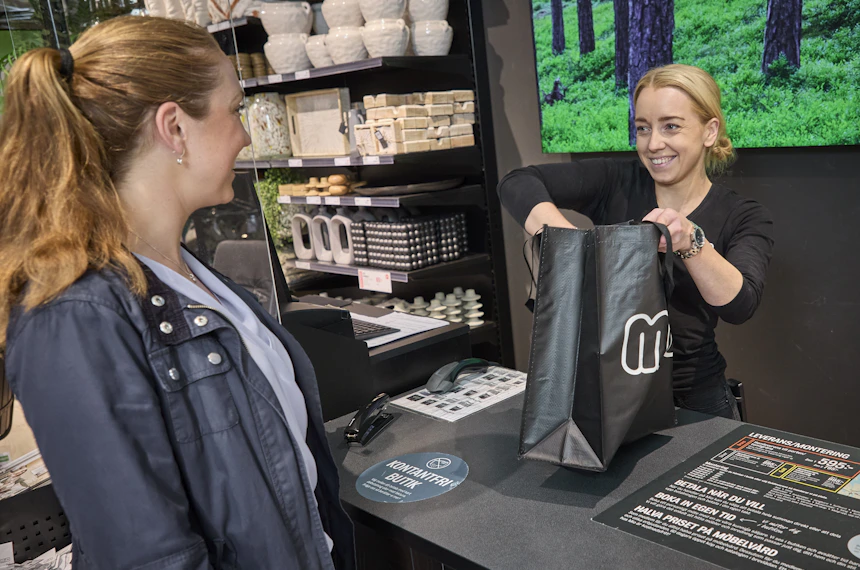 Kvinnlig butiksarbetare hjälper kund att plocka ner varor i shoppingpåse vid kassan i Miobutik.